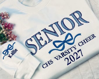 Puede incluir: Una sudadera blanca de cuello redondo con texto bordado en azul que dice SENIOR junto a un gráfico de lazo, y CHS VARSITY CHEER 2027 debajo. El puño de la manga está bordado con un lazo azul y el nombre Kallie en letras cursivas azules.