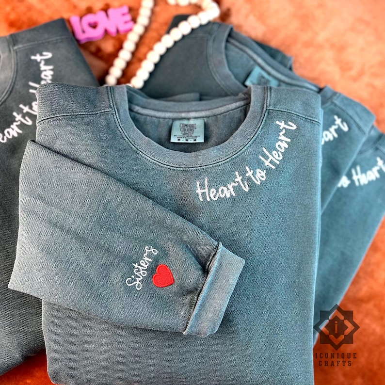 以下が含まれることがあります： 白い刺繍が施された、ターコイズブルーのトレーナーが3枚。トレーナーの前には「Heart to Heart」と刺繍されています。1枚のトレーナーには、赤いハートと「Sisters」という刺繍が袖に施されています。