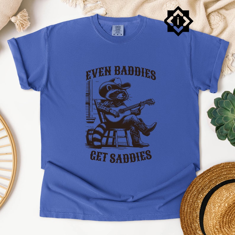 May include: A blue t-shirt with a black graphic of a raccoon wearing a cowboy hat and playing a guitar. The text "Even Baddies Get Saddles" is printed on the shirt.