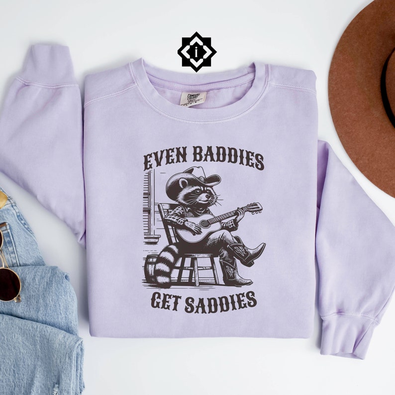 May include: A lavender sweatshirt with a black graphic of a raccoon wearing a cowboy hat and playing a guitar. The text "Even Baddies Get Saddies" is printed on the sweatshirt.