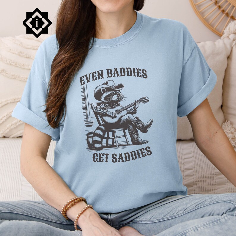 May include: A light blue t-shirt with a graphic of a raccoon wearing a cowboy hat and playing a guitar. The text "Even Baddies Get Saddles" is printed on the shirt.
