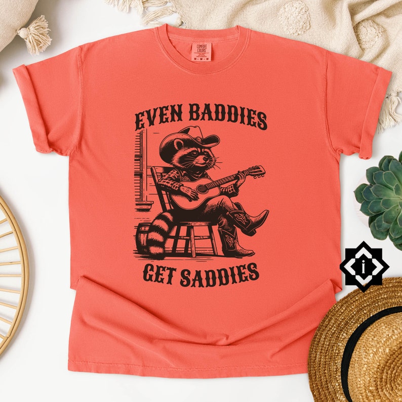 May include: A coral orange t-shirt with a black and white illustration of a raccoon wearing a cowboy hat and playing a guitar. The text "EVEN BADDIES GET SADDIES" is printed on the shirt.