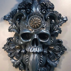 May include: A silver skull sculpture with intricate floral details and a gold centrepiece. The skull is surrounded by ornate, swirling designs.