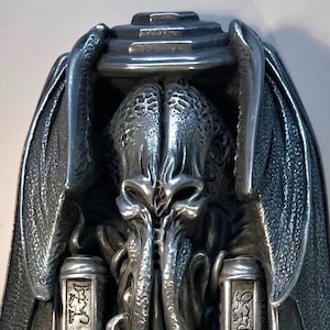 Könnte beinhalten: Eine silberfarbene Metallskulptur einer Cthulhu-ähnlichen Kreatur mit Tentakeln und Flügeln. Die Kreatur hält zwei Schriftrollen mit Text darauf. Die Skulptur ist auf einem Sockel mit einem dekorativen Design montiert.