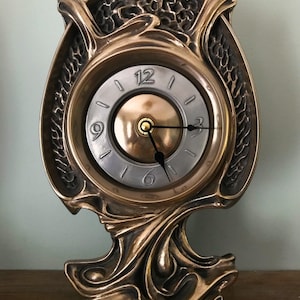 May include: An Art Nouveau style clock with a metallic bronze finish. The clock face is silver with black hands and numerals. The clock measures approximately 20 cm tall and 15 cm wide.