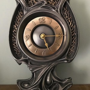 Puede incluir: Un reloj de estilo Art Nouveau con un marco de metal plateado y una esfera dorada. El reloj tiene un diseño decorativo y fluido con una esfera redonda y números romanos. Las manecillas señalan las 5:30.