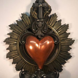 Peut inclure: Un cœur de couleur cuivre avec une auréole dorée et une croix argentée au sommet. Le cœur est entouré d'un soleil éclatant doré.