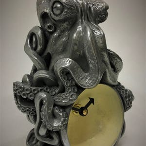 Op de afbeelding: Een grijze metalen octopusklok met een ronde, gele wijzerplaat en zwarte wijzers. De octopus is afgebeeld met gedetailleerde tentakels en een getextureerd oppervlak.