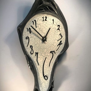 Puede incluir: Un reloj de pared de fusión negro y blanco con una cara blanca agrietada y marcadores de hora romanos negros. Las manecillas del reloj son negras.