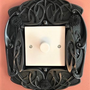 May include: A black metal light switch cover with an intricate Celtic knot design and floral accents. The switch cover has a white light switch with a single button.