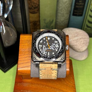 May include: A square-faced wristwatch with a black dial and silver-tone hands and markers. The watch features a world time bezel with city names. The watch has a cork band and is displayed on a wooden stand. The watch is a stylish accessory.