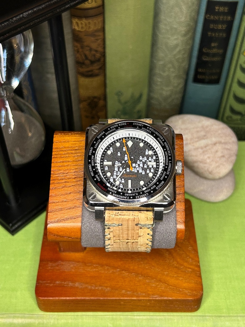 May include: A square-faced wristwatch with a black dial and silver-tone hands and markers. The watch features a world time bezel with city names. The watch has a cork band and is displayed on a wooden stand. The watch is a stylish accessory.