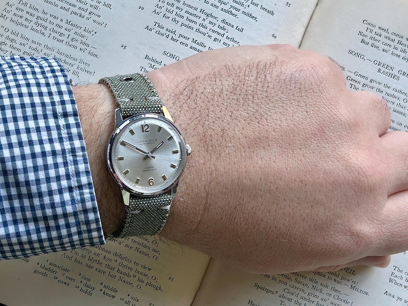 May include: A silver wristwatch with a white face and black hands. The watch has a green canvas strap with white stitching. The watch is on a person's wrist.