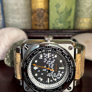May include: A square-faced wristwatch with a black dial featuring white markers and city names. The watch has an orange second hand and a cork and green canvas band. The watch face displays the word "Automatic".