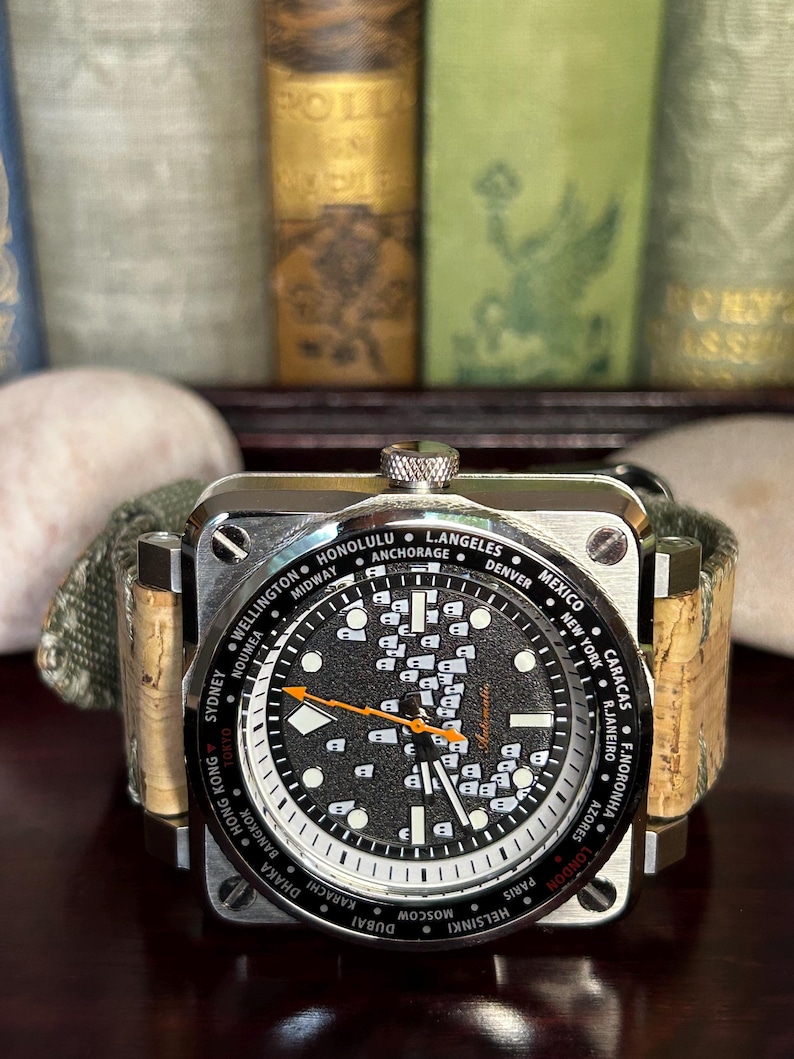 May include: A square-faced wristwatch with a black dial featuring white markers and city names. The watch has an orange second hand and a cork and green canvas band. The watch face displays the word "Automatic".