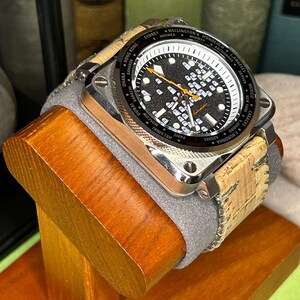 May include: A square-faced wristwatch with a black dial and white markings, including world city names. The watch has a silver-colored case and a cork and gray band. It is displayed on a wooden stand.