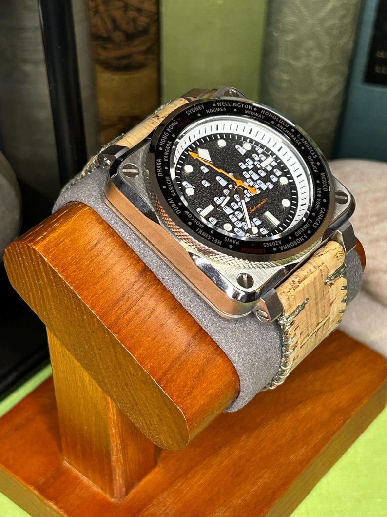 May include: A square-faced wristwatch with a black dial and white markings, including world city names. The watch has a silver-colored case and a cork and gray band. It is displayed on a wooden stand.
