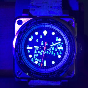 May include: A square-faced wristwatch with a dark blue face and a light blue glow. The watch face displays city names and small, white, glowing shapes. The watch has a brown leather band and a metal case.