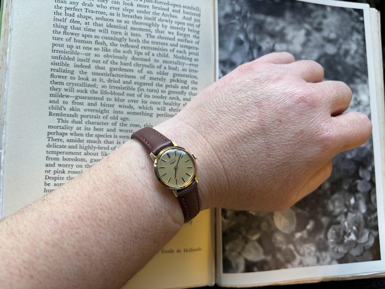 May include: A gold-toned wristwatch with a brown leather band is displayed. The watch has a round face with gold hands and markers. The watch is on a wrist, with an open book in the background.