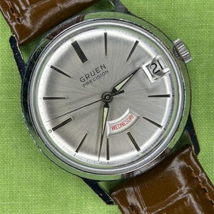 May include: A vintage Gruen Precision wristwatch with a silver dial, black markers, and a date display. The watch has a stainless steel case and a brown leather strap. The word "Wednesday" is visible on the dial.