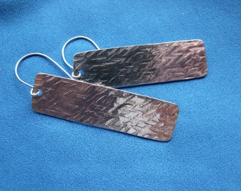 May include: A pair of silver rectangular earrings with a textured surface. The earrings hang from silver hooks and are displayed against a blue background. The earrings have a hammered metal appearance.