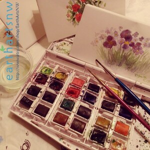 May include: A white plastic watercolor palette with 24 paint wells, filled with various colors of watercolor paint. Two paintbrushes are on top of the palette, one red and one blue. The palette is on a white surface.