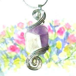 May include: A silver wire wrapped pendant featuring a purple and white amethyst gemstone. The pendant is hanging from a silver chain against a blurred floral background.