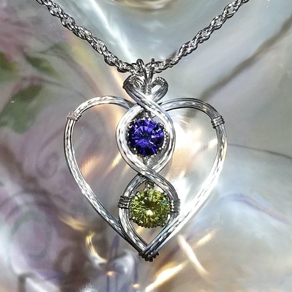 Puede incluir: Un collar con un colgante de corazón de alambre plateado con una piedra preciosa púrpura y verde. El colgante presenta un diseño de doble corazón con las piedras preciosas engastadas en el centro.