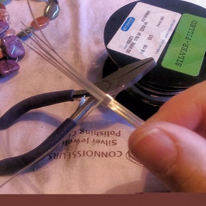 May include: A close-up of a hand holding a spool of silver-filled wire. The wire is being cut with a pair of pliers. The spool is labeled "Silver-Filled".