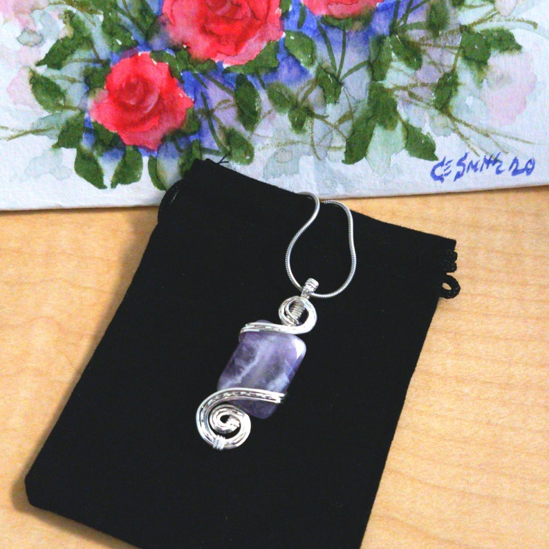May include: A silver wire wrapped pendant featuring a purple amethyst stone. The pendant is displayed on a black velvet pouch.