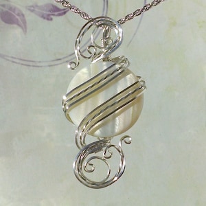 May include: A silver wire wrapped pendant featuring a white mother of pearl cabochon. The pendant has a delicate, swirling design.