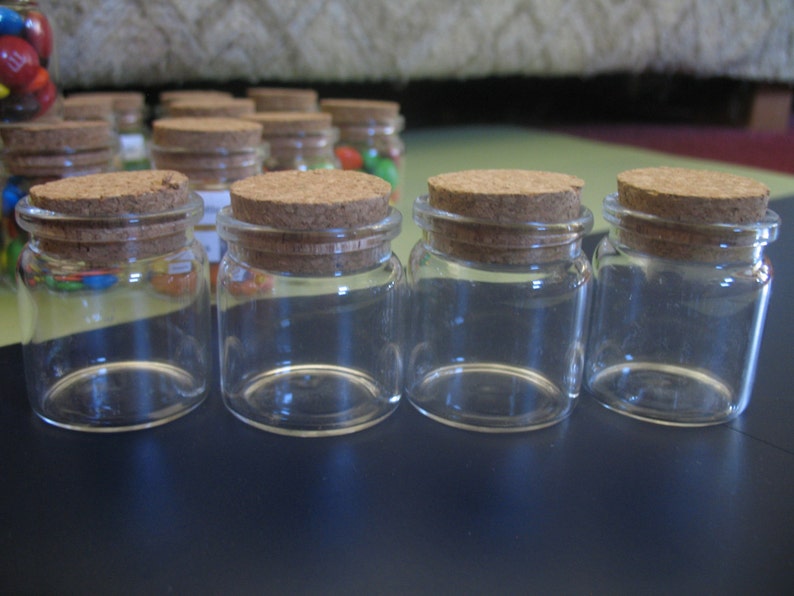 May include: Four clear glass jars with cork lids. The jars are empty and have a round shape.