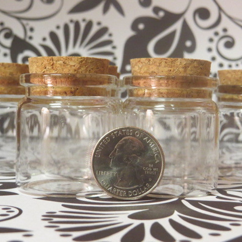 May include: Four clear glass jars with cork lids. The jars are small, about the size of a quarter. The jars are on a black and white patterned surface.