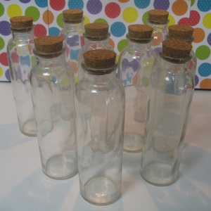 May include: Ten clear glass bottles with cork stoppers. The bottles are arranged in a semi-circle on a white surface.