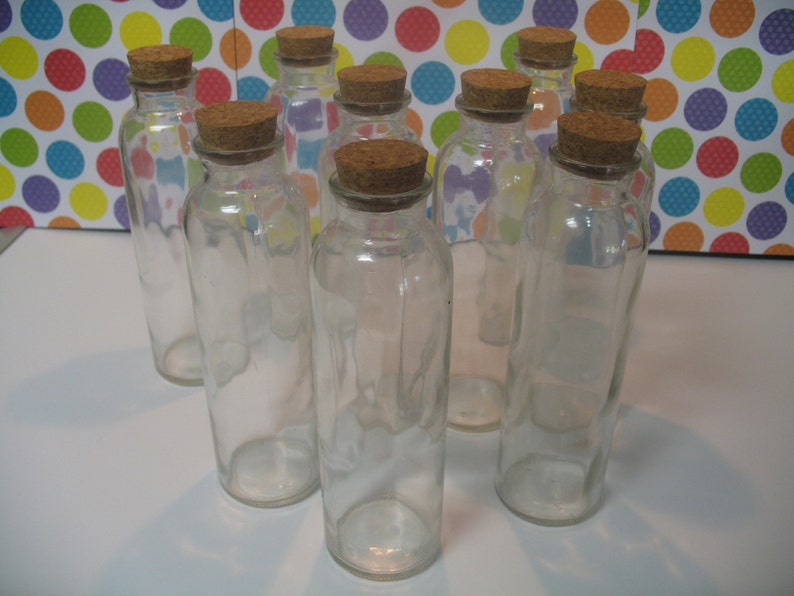 May include: Ten clear glass bottles with cork stoppers. The bottles are arranged in a semi-circle on a white surface.