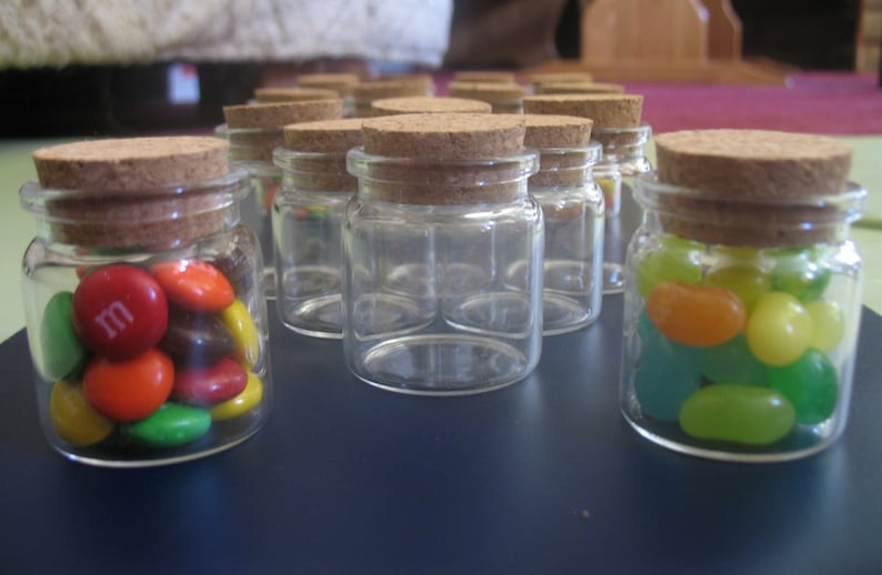 May include: A group of small glass jars with cork lids filled with colorful candies. The jars are arranged in a row on a dark blue surface. Some jars contain M&M's candies, while others contain jelly beans.
