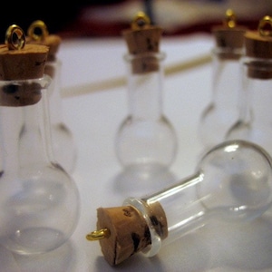 Pode incluir: Cinco garrafas de vidro transparente com rolhas de cortiça e tampas de metal dourado. As garrafas são redondas com um pescoço pequeno e estão dispostas em grupo sobre uma superfície branca.