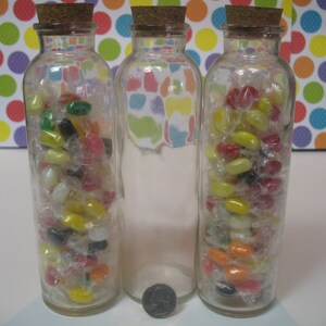 May include: Three clear glass bottles with cork tops filled with colorful jelly beans. The bottles are arranged in a row on a white surface.