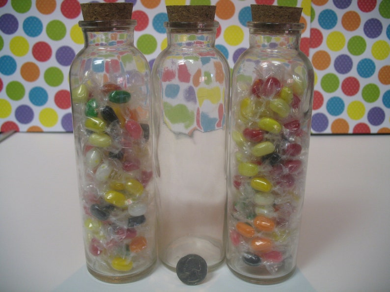 May include: Three clear glass bottles with cork tops filled with colorful jelly beans. The bottles are arranged in a row on a white surface.