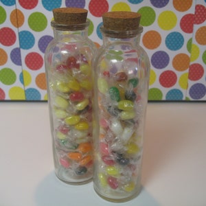 May include: Two clear glass bottles filled with colorful jelly beans. The bottles have cork stoppers and are filled with a variety of colors, including red, yellow, green, and white.