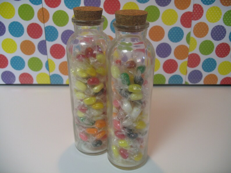 May include: Two clear glass bottles filled with colorful jelly beans. The bottles have cork stoppers and are filled with a variety of colors, including red, yellow, green, and white.