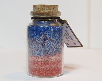 Puede incluir: Un pequeño frasco de vidrio con tapa de corcho lleno de purpurina roja, blanca y azul. Una etiqueta adjunta al frasco dice "Galaxy in a Jar".