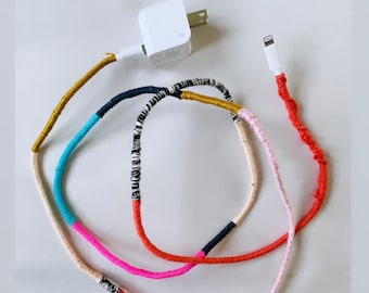 May include: A white charging brick and a white iPhone charging cable wrapped in colorful yarn. The yarn is red, pink, blue, black, white, and gold.