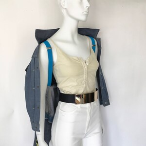 K&ouml;nnte beinhalten: Ein wei&szlig;es Tanktop mit Knopfleiste, getragen mit einer blauen Jeansjacke und einer wei&szlig;en Hose. Ein schwarzer G&uuml;rtel mit goldener Schnalle wird &uuml;ber der Hose getragen. Blaue Hosentr&auml;ger werden &uuml;ber dem Tanktop getragen.