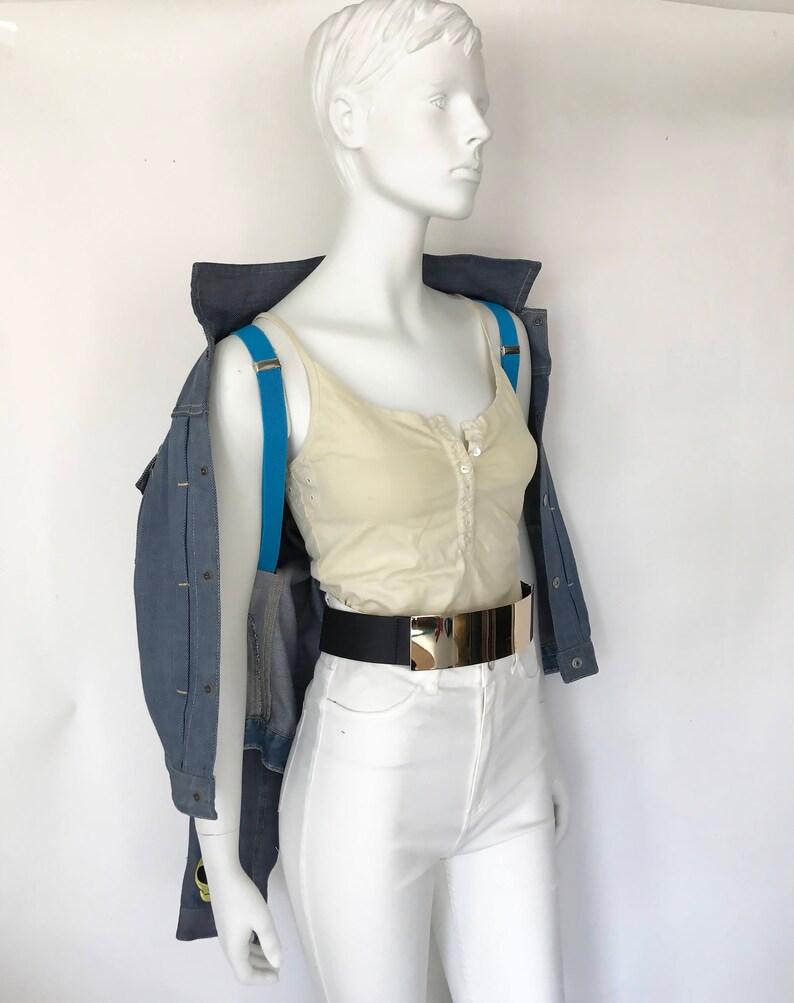 K&ouml;nnte beinhalten: Ein wei&szlig;es Tanktop mit Knopfleiste, getragen mit einer blauen Jeansjacke und einer wei&szlig;en Hose. Ein schwarzer G&uuml;rtel mit goldener Schnalle wird &uuml;ber der Hose getragen. Blaue Hosentr&auml;ger werden &uuml;ber dem Tanktop getragen.