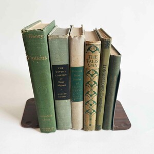May include: A collection of antique books in various shades of green and beige, held upright by a brown bookend. The book spines display titles like "Henry Options" and "The Divine Comedy". The books are arranged on a white surface.