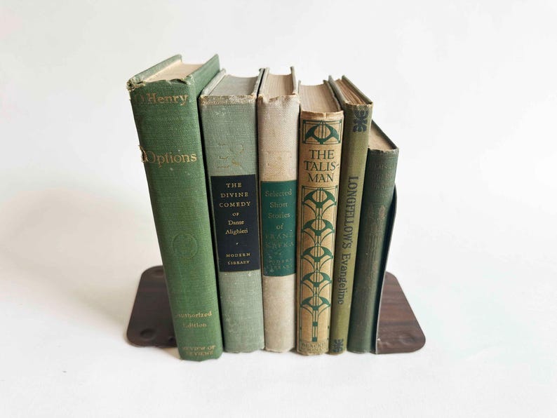 May include: A collection of antique books in various shades of green and beige, held upright by a brown bookend. The book spines display titles like "Henry Options" and "The Divine Comedy". The books are arranged on a white surface.