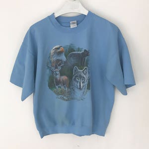 May include: A pale blue short-sleeved sweatshirt with a wildlife design. The design features an eagle, a deer, a wolf, and a bear. The sweatshirt is suspended on a white hanger against a white backdrop.