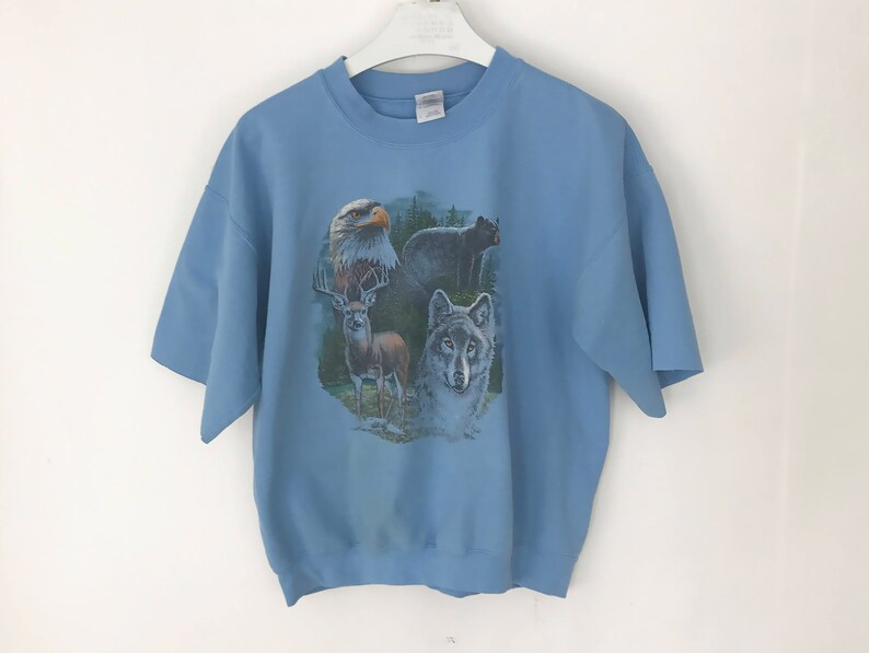 May include: A pale blue short-sleeved sweatshirt with a wildlife design. The design features an eagle, a deer, a wolf, and a bear. The sweatshirt is suspended on a white hanger against a white backdrop.