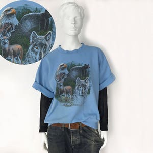 May include: A light blue t-shirt with a wildlife graphic depicting an eagle, bear, deer, and wolf. The sleeves are rolled up, and it's worn over a black long-sleeved top. The mannequin is wearing blue jeans and a brown belt.